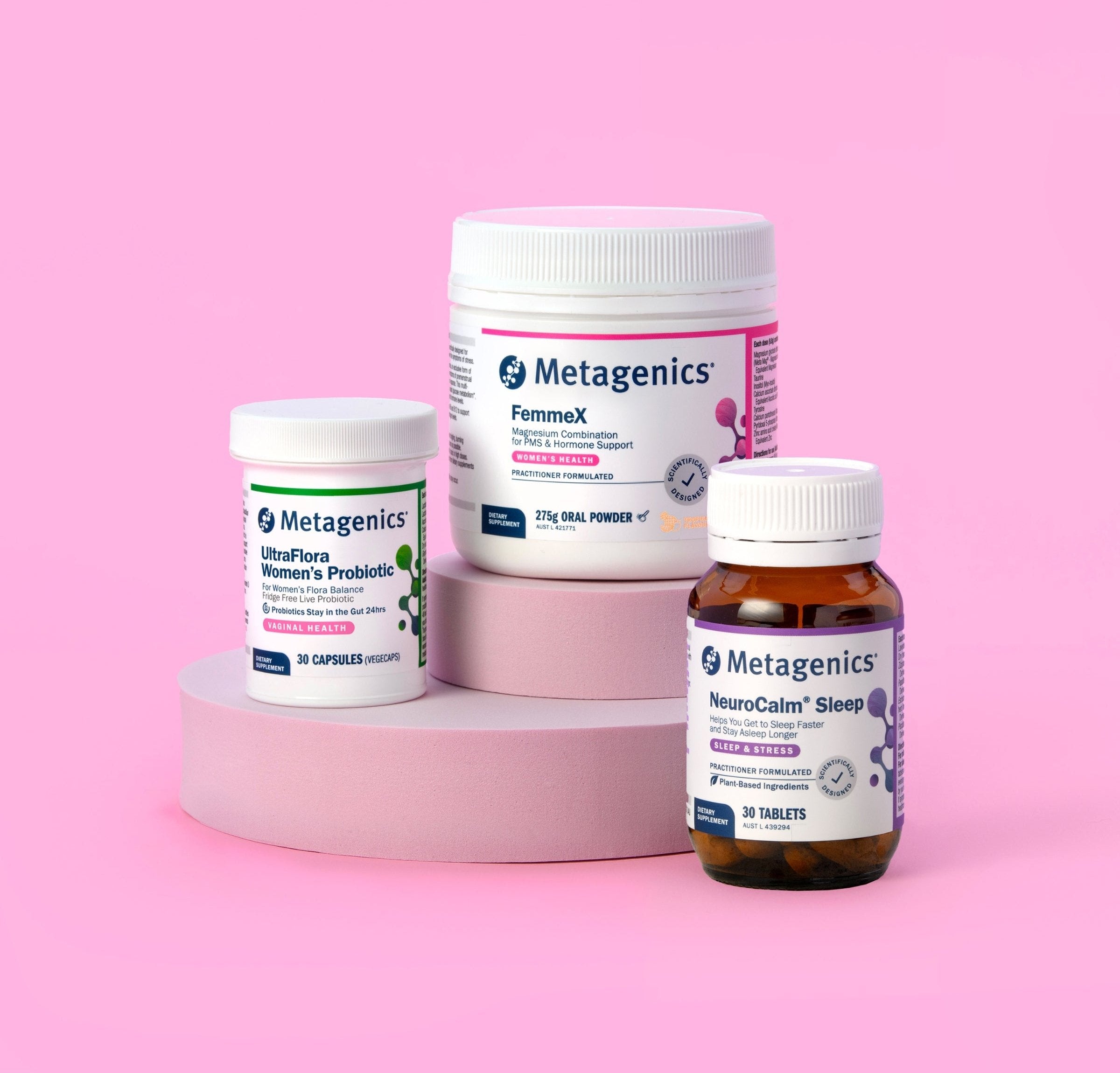 Three Metagenics supplement bottles displayed on pastel pink cylindrical platforms against a pink background. The products include 'UltraFlora Women's Probiotic', 'FemmeX' and 'NeuroCalm Sleep'.